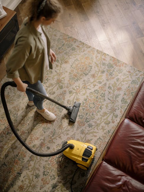 A person using a vacuum cleaner to perform deep cleaning on a dark carpeted floor in an indoor space with modern architectural design, illuminated by overhead lights. The individual is dressed in ligh