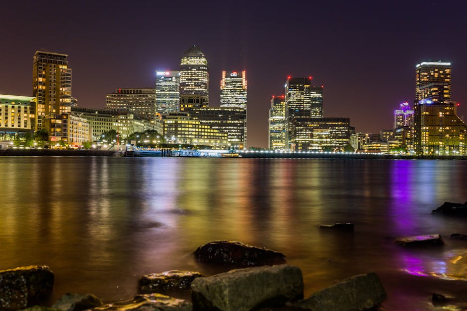 A cityscape view along the River Thames in Canary Wharf, featuring modern office buildings with glass facades reflecting the sky and surrounding structures. A red London Underground train crosses an e