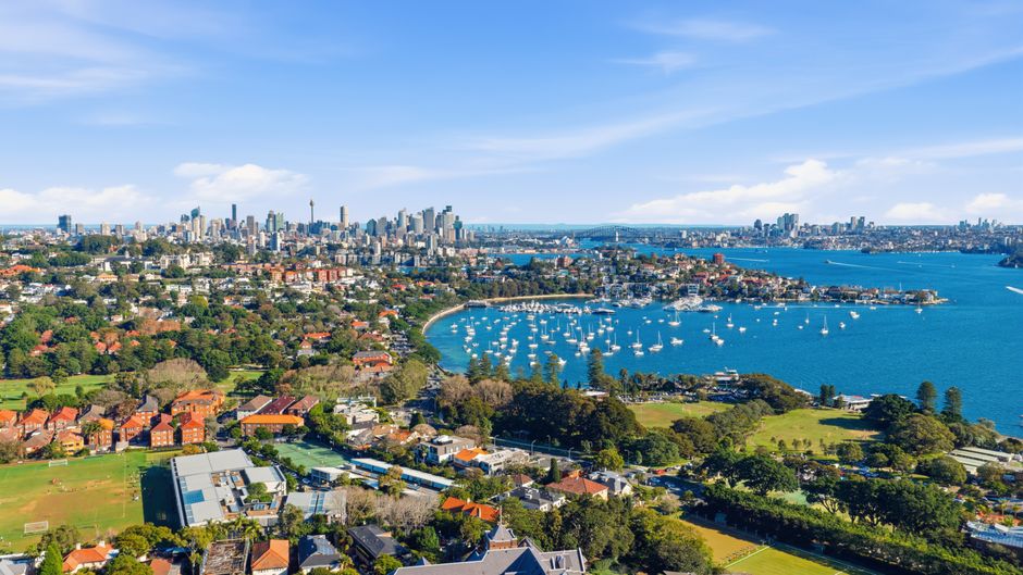 Aerial view of a residential and commercial area with a large body of water featuring numerous sailboats anchored near the shore. In the foreground, there are green parks, sports fields, and rows of h