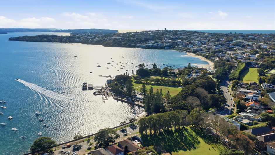 Aerial view of a residential and commercial area with a large body of water featuring numerous sailboats anchored near the shore. In the foreground, there are green parks, sports fields, and rows of h